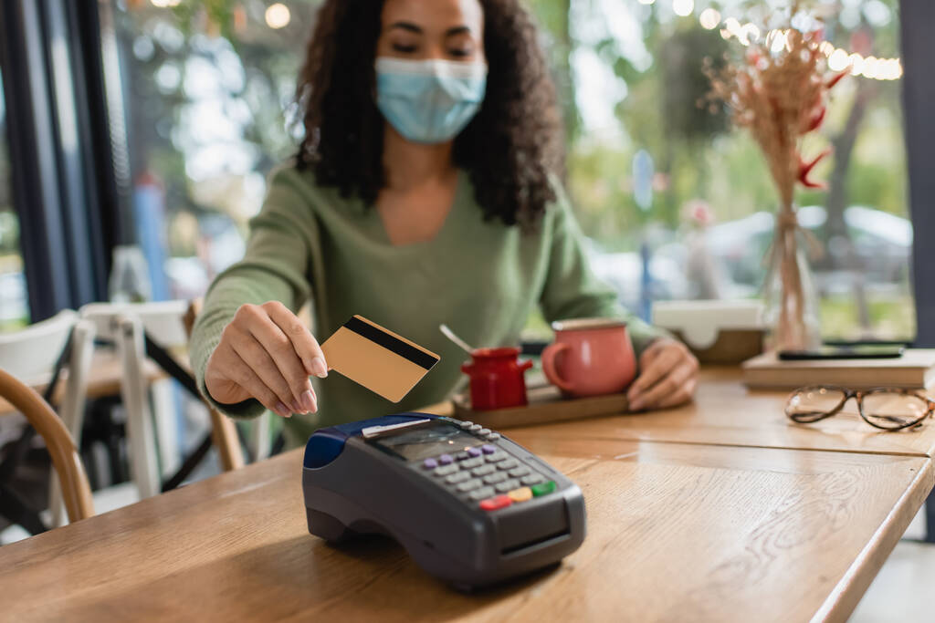 кредитная карта возле считывателя кредитных карт в руке африканской американки в медицинской маске на размытом фоне  - Фото, изображение