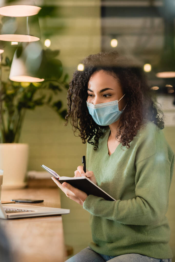 Afrikanerin in medizinischer Maske mit Notizbuch und Stift beim Blick auf Laptop  - Foto, Bild