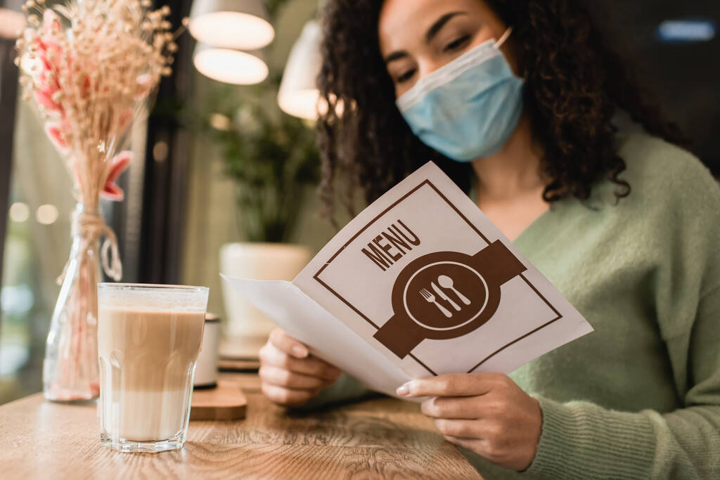 Afrikanerin in medizinischer Maske liest Menü in der Nähe von Glas Latte im Café  - Foto, Bild