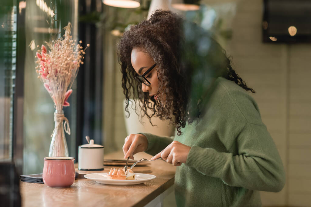 Afrikanerin in Brille hält Besteck in der Nähe von Torte auf Teller - Foto, Bild