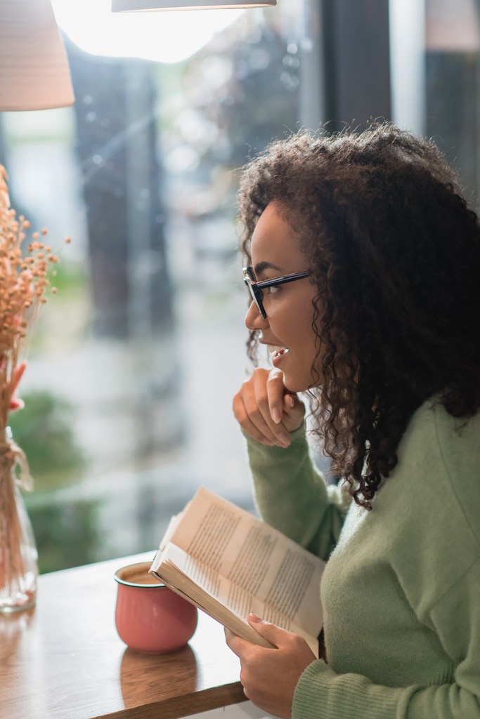 vista laterale della donna afro-americana positiva negli occhiali da vista lettura libro in caffè - Foto, immagini