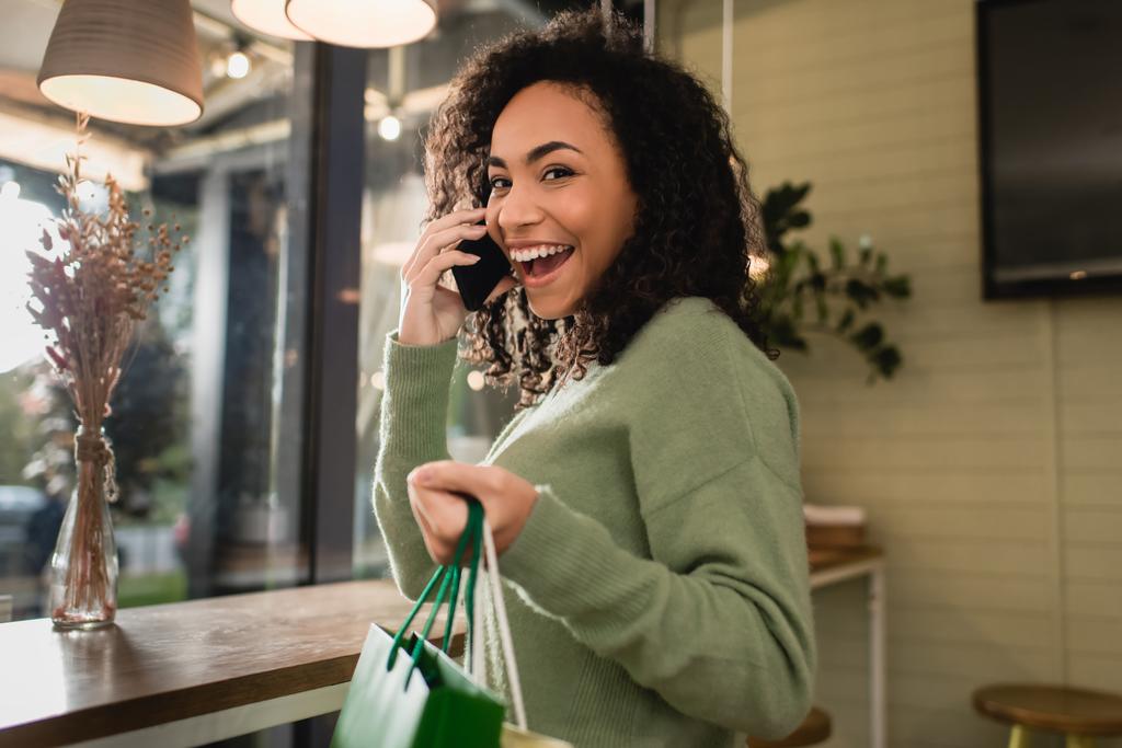lächelnde Afroamerikanerin spricht auf Smartphone und hält Einkaufstüten im Café - Foto, Bild