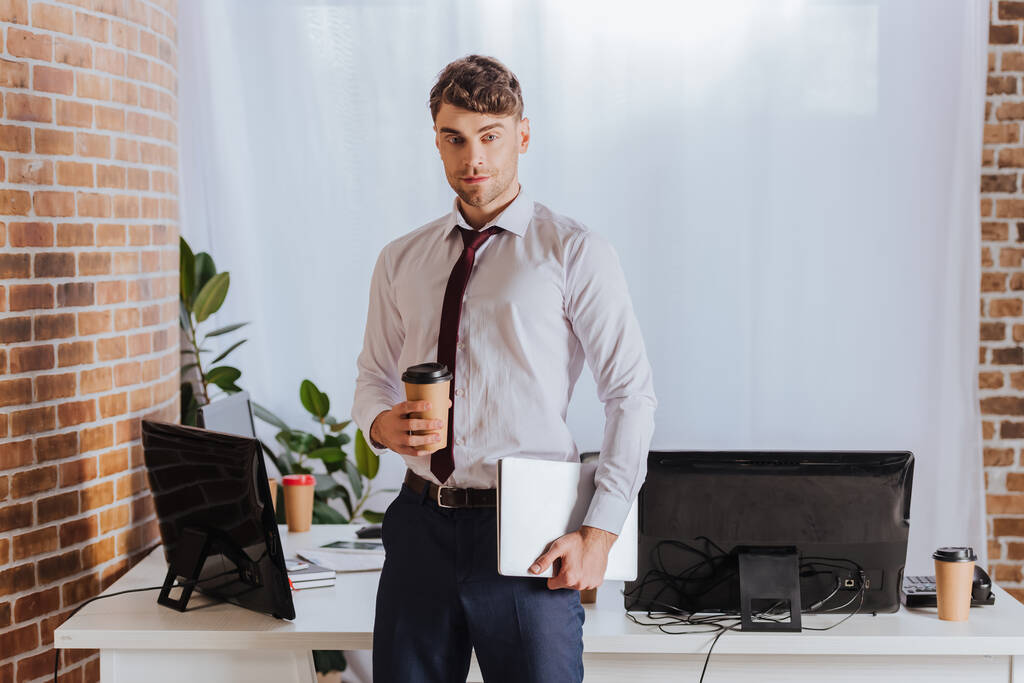 Młody biznesmen trzyma kawę na wynos i laptopa w pobliżu komputerów w biurze  - Zdjęcie, obraz