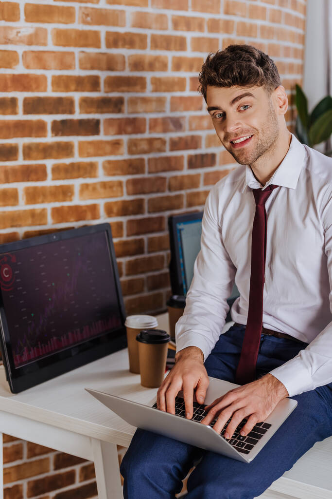 Empresário sorrindo usando laptop perto de computador com gráficos e café para ir em fundo turvo  - Foto, Imagem
