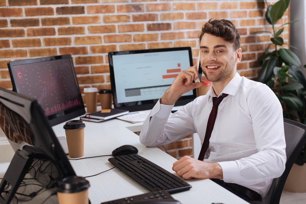 Hombre de negocios sonriente hablando en un teléfono inteligente cerca de computadoras con gráficos financieros y café para llevar en un fondo borroso  - Foto, Imagen