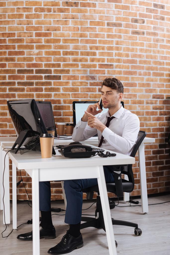 Young businessman pointing with finger at computer while talking on smartphone near takeaway coffee  - Photo, Image