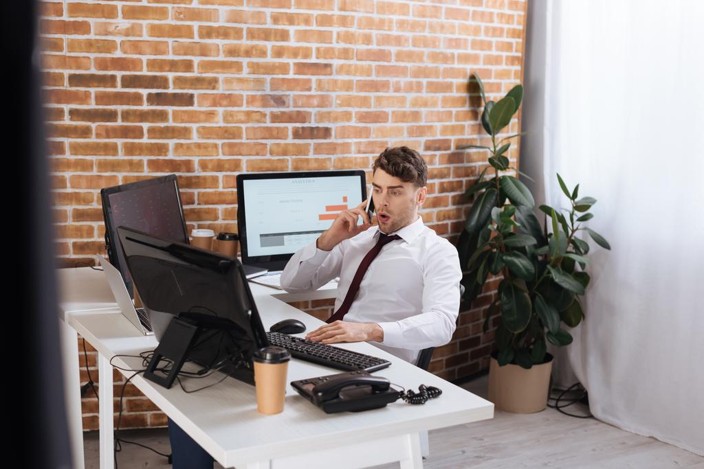 Hombre de negocios sorprendido hablando en el teléfono inteligente mientras revisa las acciones financieras en las computadoras  - Foto, imagen