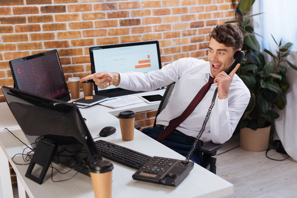 Cheerful businessman talking on telephone and pointing with finger at computer near coffee to go on blurred foreground  - Photo, Image