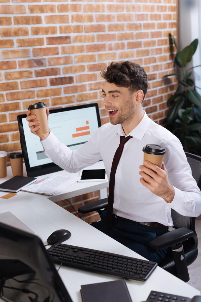 前景にぼやけたコンピュータの近くに座っているテイクアウトコーヒーと肯定的なビジネスマン  - 写真・画像