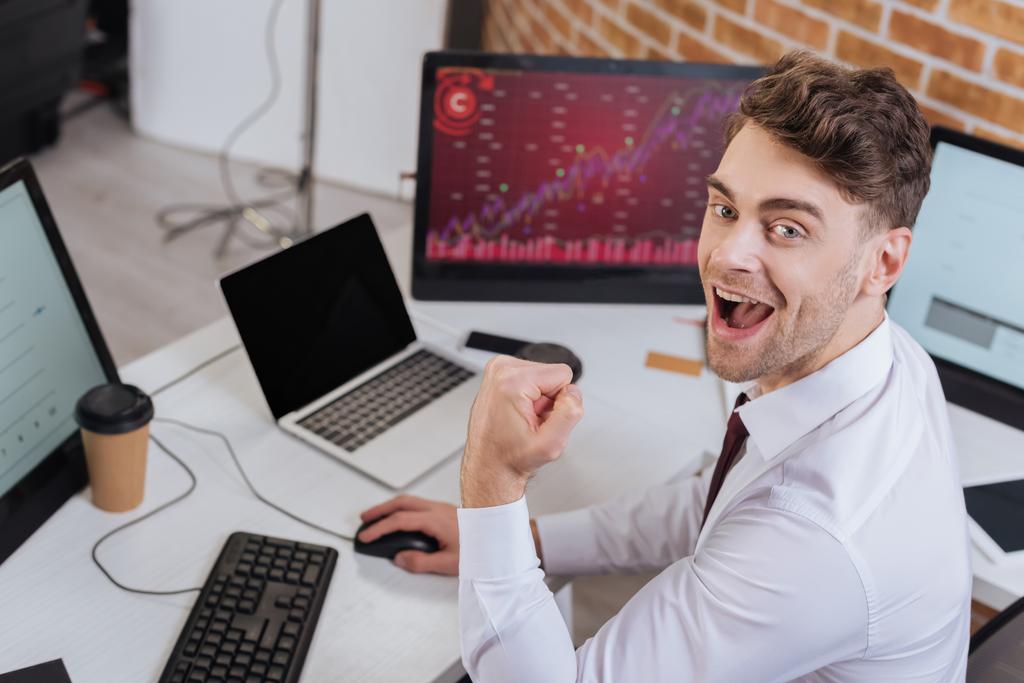 Izgatott üzletember, aki kamerába néz, miközben igen gesztust mutat, és ellenőrzi a pénzügyi kurzusokat a számítógépen homályos háttérrel  - Fotó, kép