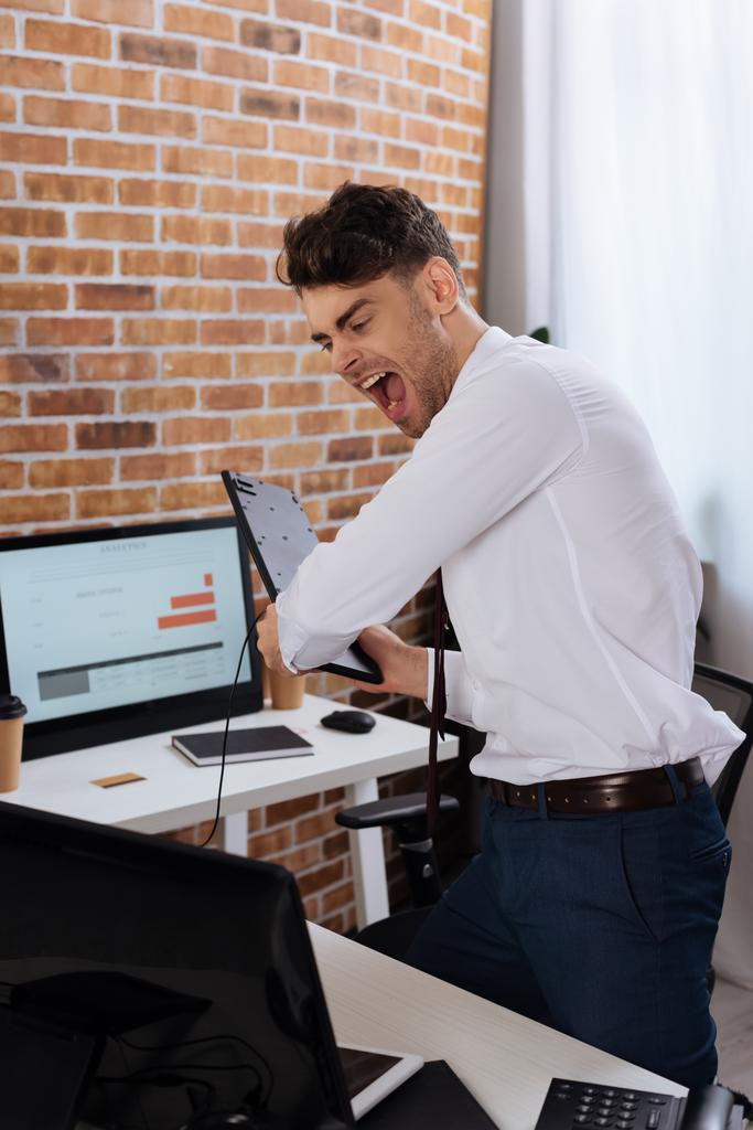 Szalony biznesmen trzymający klawiaturę i krzyczący w pobliżu komputera na niewyraźnym pierwszym planie  - Zdjęcie, obraz