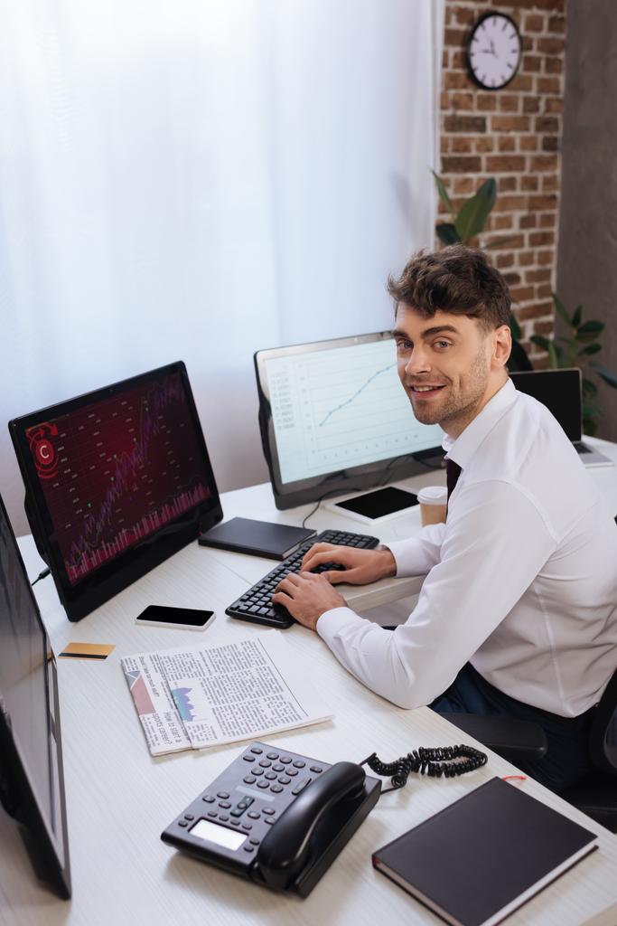 Uomo d'affari sorridente che utilizza il computer con grafici sul monitor vicino al telefono, giornali e notebook in primo piano sfocato  - Foto, immagini