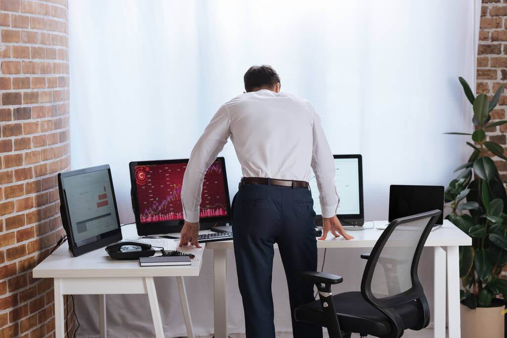 Вид ззаду бізнесмена, що стоїть біля комп'ютерів з графіками в офісі
  - Фото, зображення