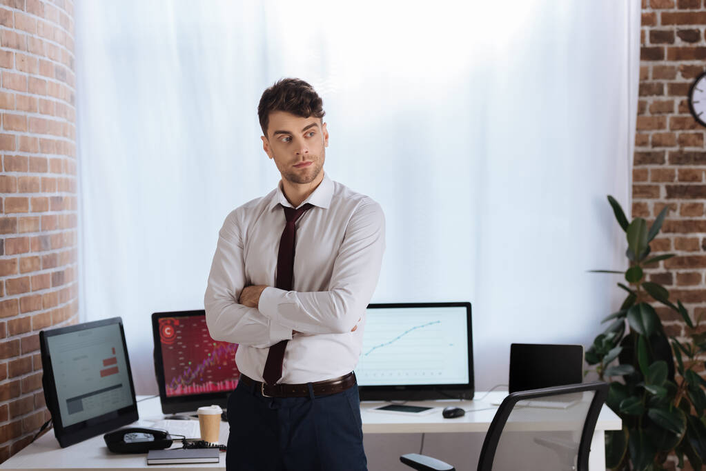 επιχειρηματίας στέκεται με σταυρωμένα τα χέρια κοντά σε υπολογιστές σε θολή φόντο στο γραφείο  - Φωτογραφία, εικόνα