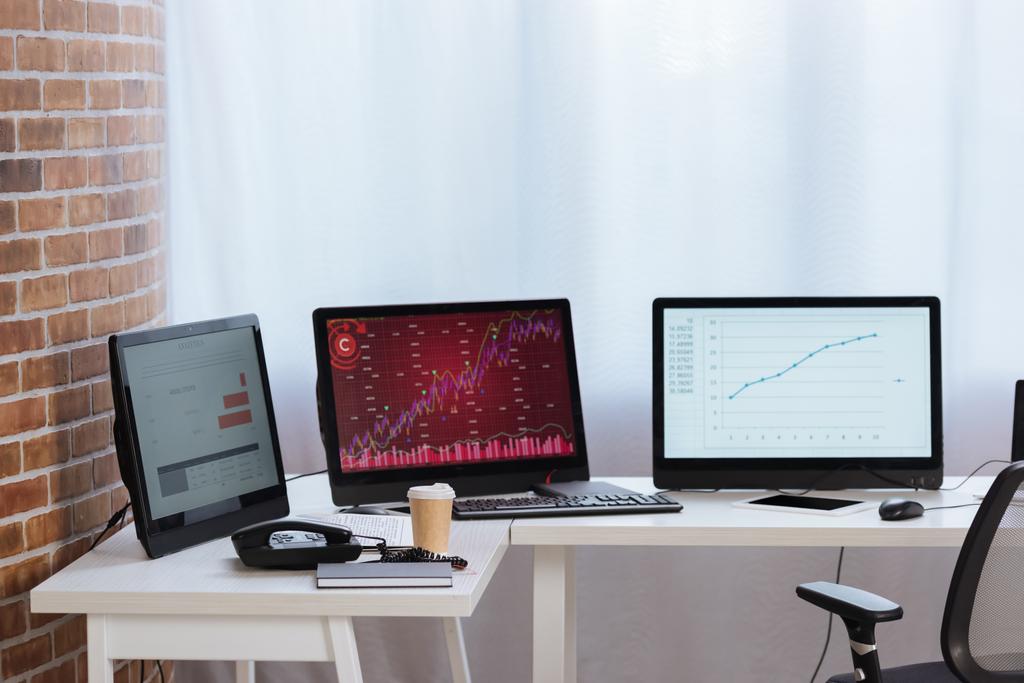 Υπολογιστές με διαγράμματα των οικονομικών αποθεμάτων στις οθόνες, καφέ για να πάει και τηλέφωνο στο γραφείο  - Φωτογραφία, εικόνα