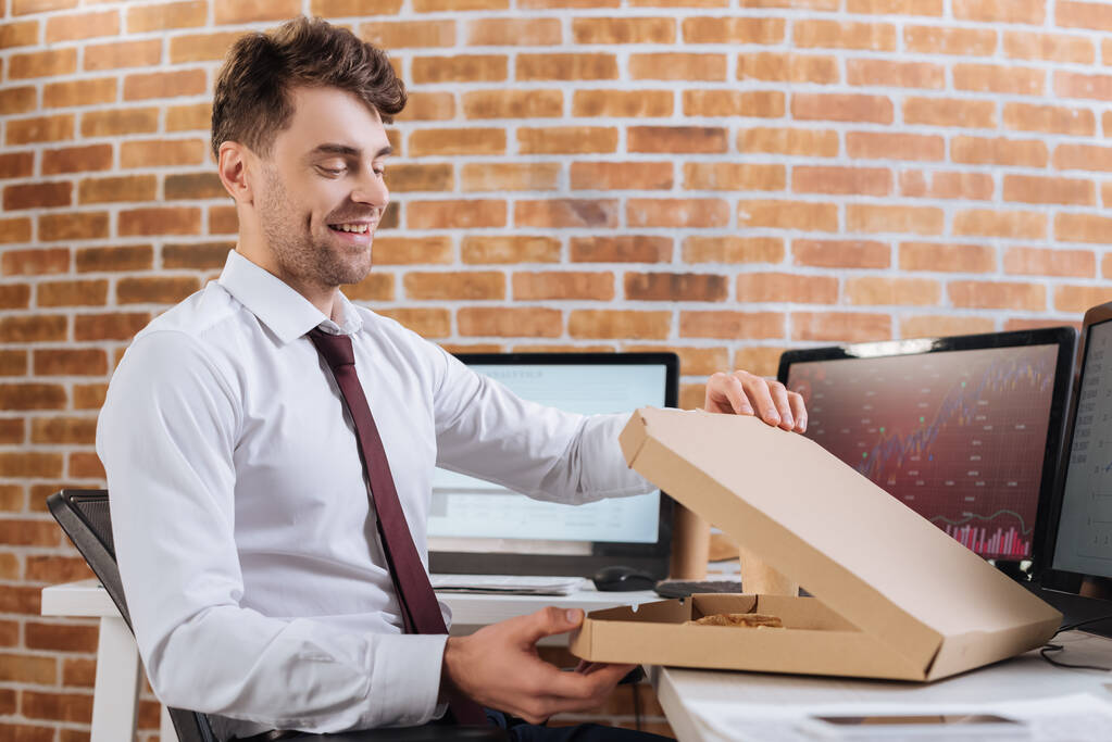 Hombre de negocios sonriente abriendo una caja de pizza cerca de computadoras en un fondo borroso  - Foto, Imagen