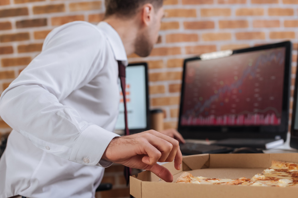 Geschäftsmann nimmt leckere Pizza in Schachtel in der Nähe von Computern auf verschwommenem Hintergrund im Büro  - Foto, Bild
