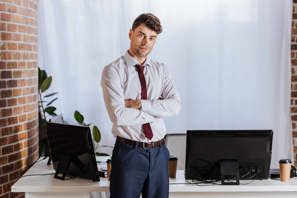 Homme d'affaires debout avec les bras croisés près des ordinateurs dans le bureau  - Photo, image