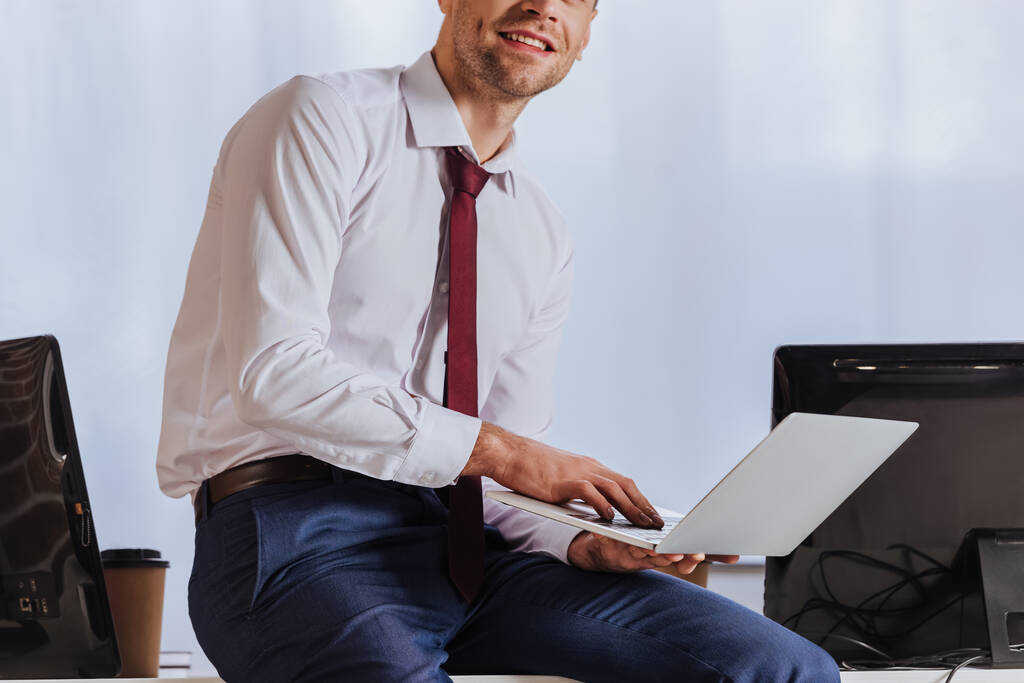 Обрезанный вид улыбающегося бизнесмена с помощью ноутбука рядом с компьютерами в офисе  - Фото, изображение