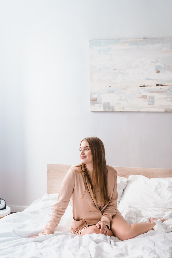 задоволена молода жінка дивиться геть, сидячи на ліжку в сучасній спальні
  - Фото, зображення