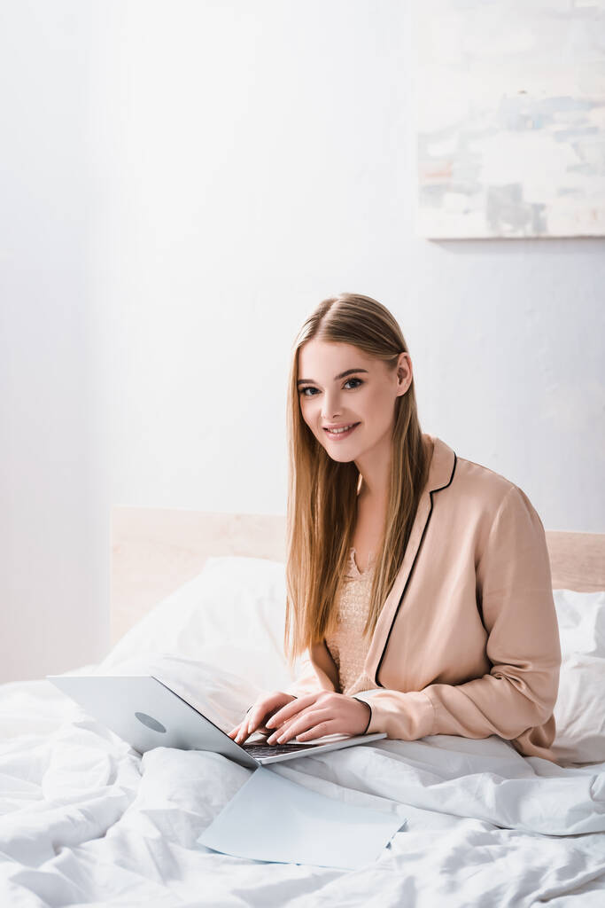 alegre joven freelancer en bata de seda utilizando el ordenador portátil en el dormitorio - Foto, Imagen