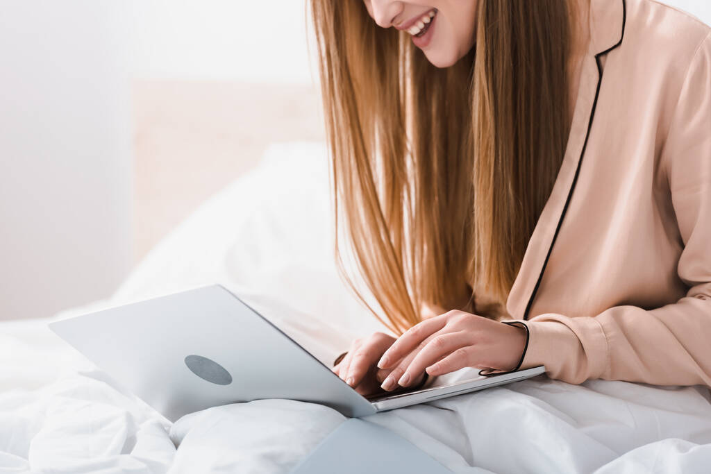 Ausgeschnittene Ansicht einer lächelnden jungen Freiberuflerin im Seidenmantel mit Laptop im Schlafzimmer - Foto, Bild
