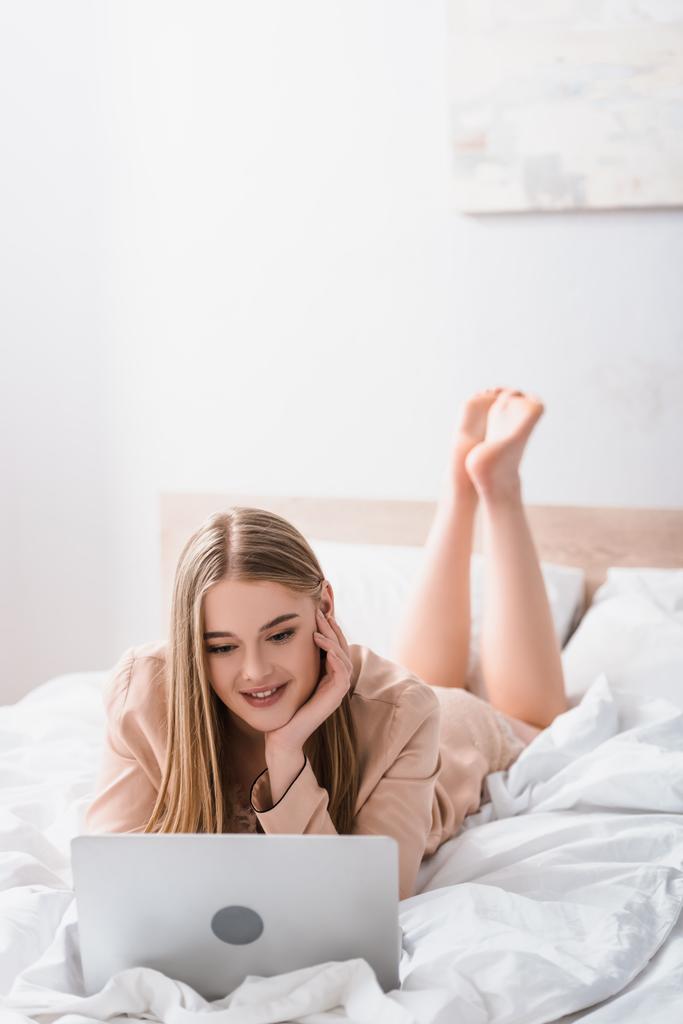 jeune pigiste en robe de soie couchée sur le lit et utilisant un ordinateur portable dans la chambre - Photo, image