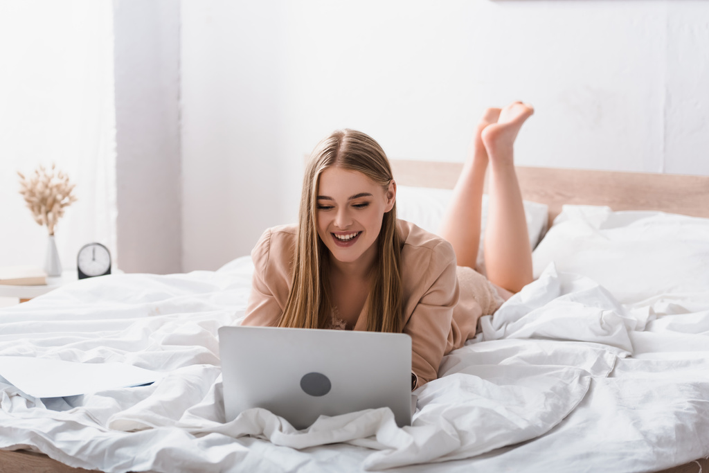 giovane freelance sorridente in abito di seta sdraiato sul letto e utilizzando il computer portatile - Foto, immagini