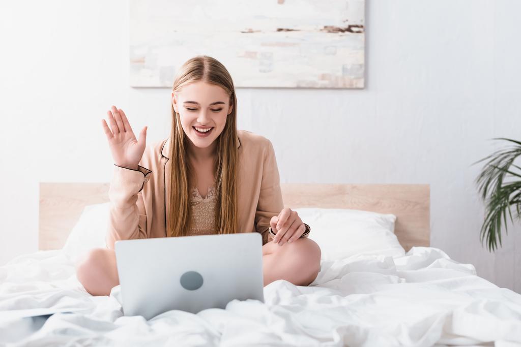 щаслива жінка в атласному халаті махає рукою, маючи відеочат на ноутбуці в спальні
 - Фото, зображення