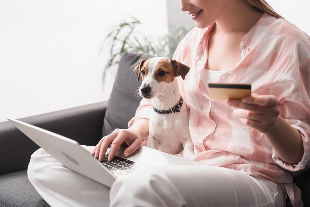 oříznutý pohled na šťastnou ženu držící kreditní kartu v blízkosti psa a notebooku při online nakupování doma - Fotografie, Obrázek