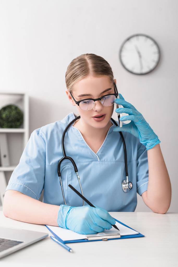 young veterinarian in glasses and latex gloves talking on smartphone near laptop on desk - Photo, Image