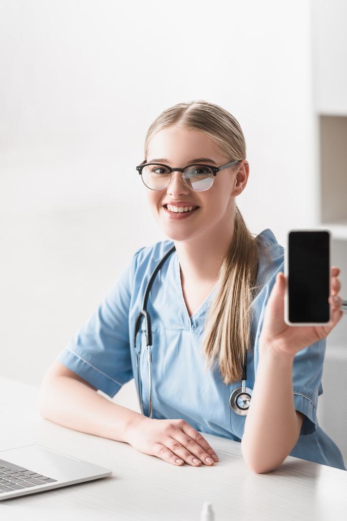 veterinário alegre em óculos segurando smartphone com tela em branco perto de laptop na mesa - Foto, Imagem