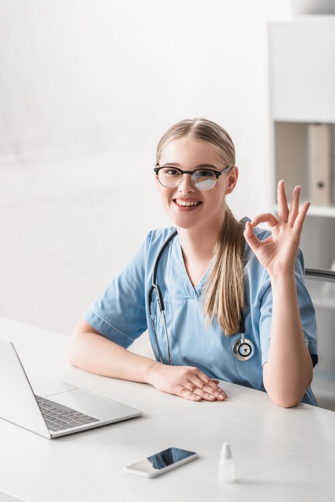 Fröhlicher Tierarzt in Brille zeigt Ok-Zeichen in der Nähe von Gadgets auf dem Tisch - Foto, Bild