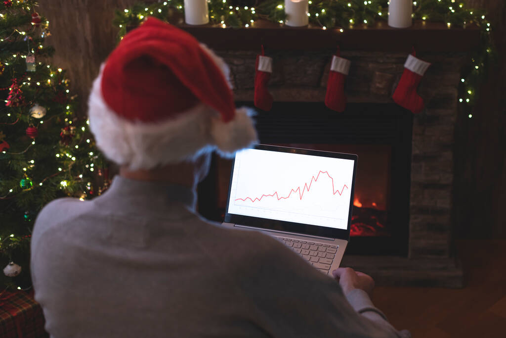 Mann mit Weihnachtsmann-Hut arbeitet am Laptop mit Grafiken und Diagrammen auf dem Bildschirm und sitzt neben Weihnachtsbaum und Kamin. - Foto, Bild