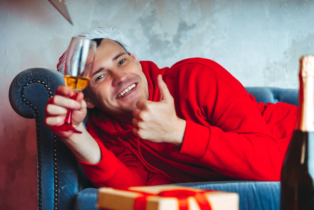 Fiatalember télapó kalapban, egy pohár pezsgővel a kanapén. Felnőtt férfi pihen a szobában. Fogalom a karácsonyi ünnepség otthon - Fotó, kép