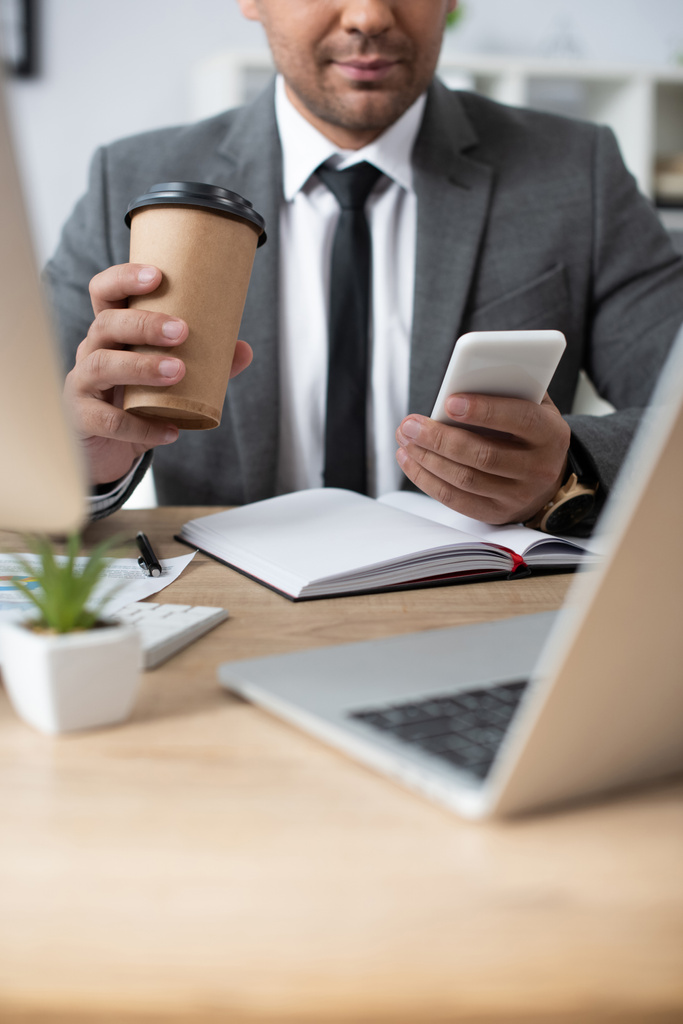 bijgesneden weergave van handelaar houden van koffie te gaan, terwijl berichten op smartphone in de buurt van laptop op wazig voorgrond - Foto, afbeelding