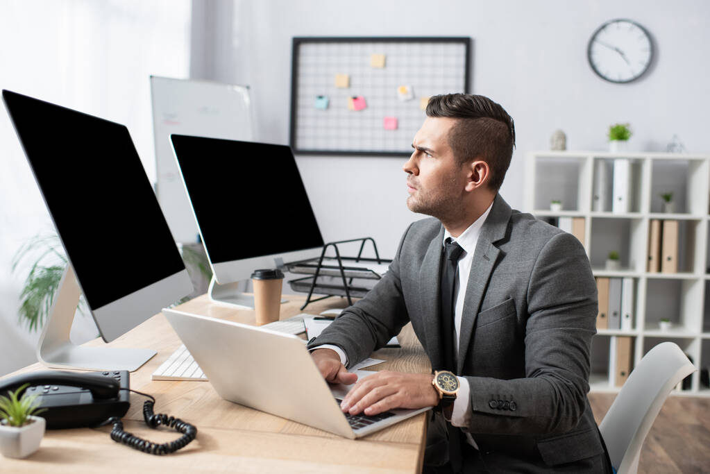 comerciante concentrado trabalhando no laptop perto de monitores com tela em branco - Foto, Imagem