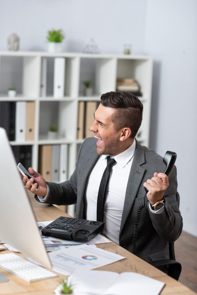 comerciante animado gritando enquanto segurando aparelho e smartphone no local de trabalho, foreground borrado - Foto, Imagem