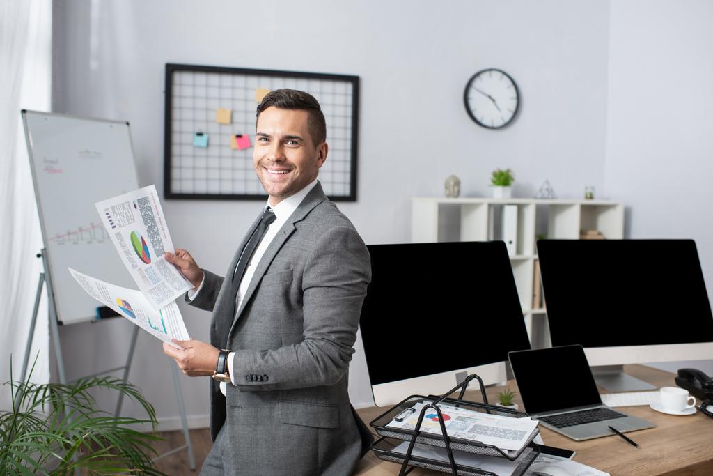 χαμογελαστός έμπορος κρατώντας infographics, ενώ στέκεται κοντά οθόνες με κενή οθόνη - Φωτογραφία, εικόνα