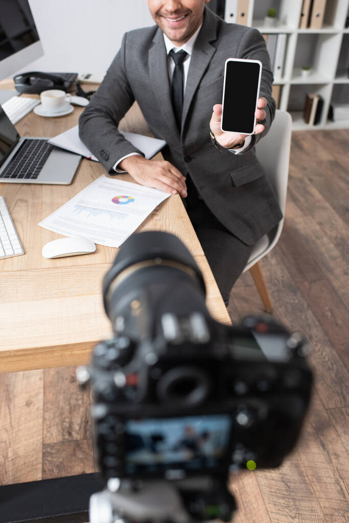 vista cortada de empresário mostrando telefone celular com tela em branco perto da câmera digital em primeiro plano borrado - Foto, Imagem