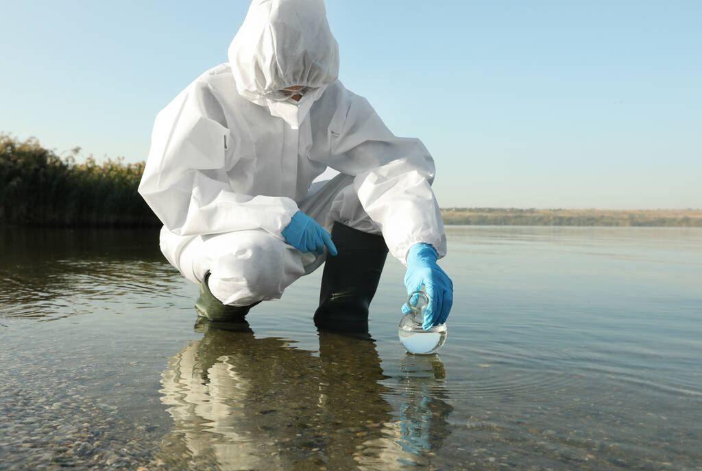 Kemiallisessa suojapuvussa oleva tiedemies, joka ottaa näytteen joesta Florence-pullolla analysoitavaksi - Valokuva, kuva
