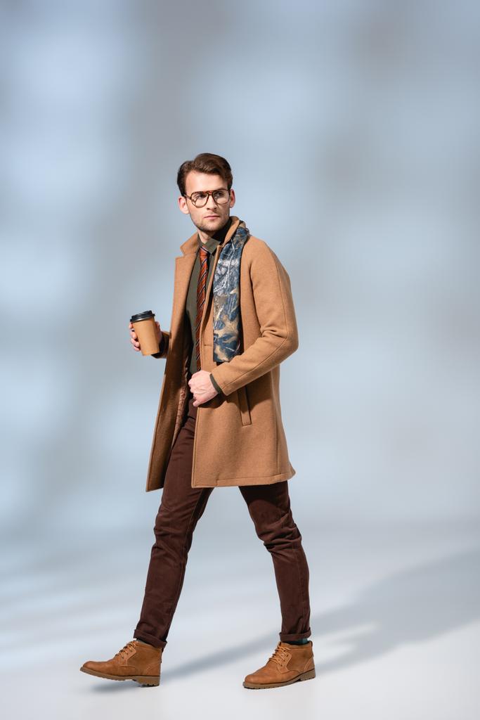 冬のコートを着たスタイリッシュな男がコーヒーを持ってグレーを歩き  - 写真・画像