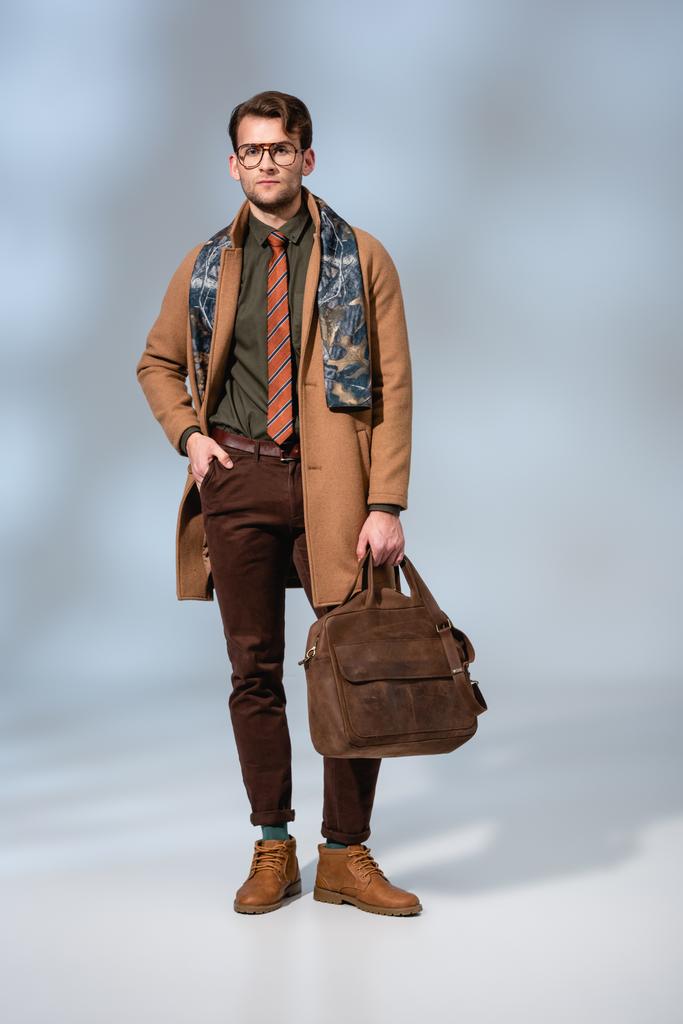 Kışlık ceketli, deri çantalı ve eldivenli, cebinde gri bir çantayla poz veren şık bir adam.  - Fotoğraf, Görsel