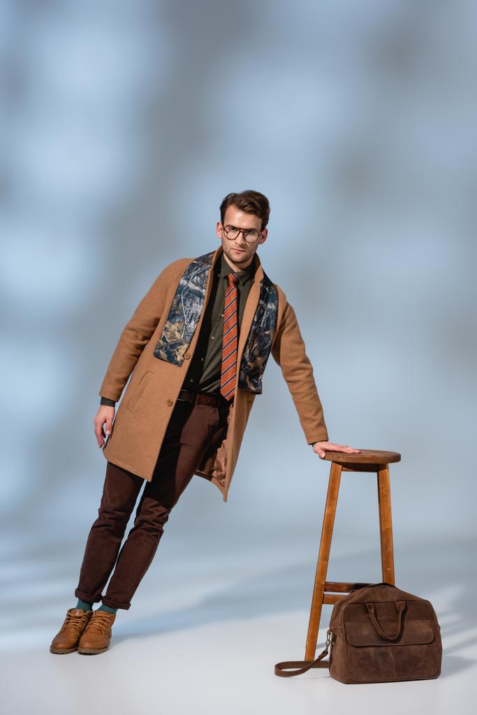 полная длина модный человек в зимнем наряде и очки опираясь на деревянный стул возле портфеля на серый - Фото, изображение
