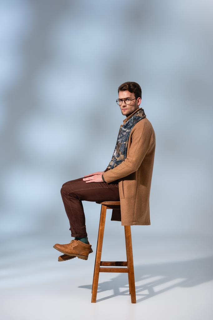 full length of stylish man in winter outfit and glasses sitting on wooden chair on grey - Photo, Image
