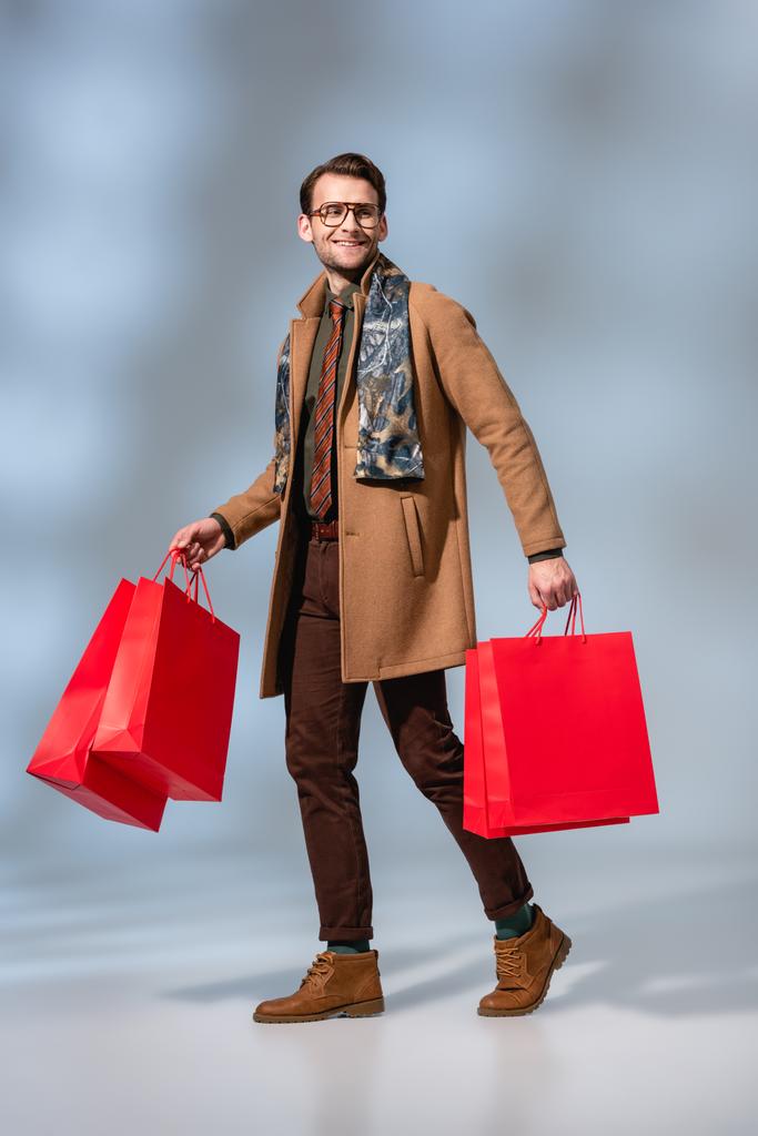 plná délka spokojený zákazník v brýlích drží papírové tašky na šedé - Fotografie, Obrázek