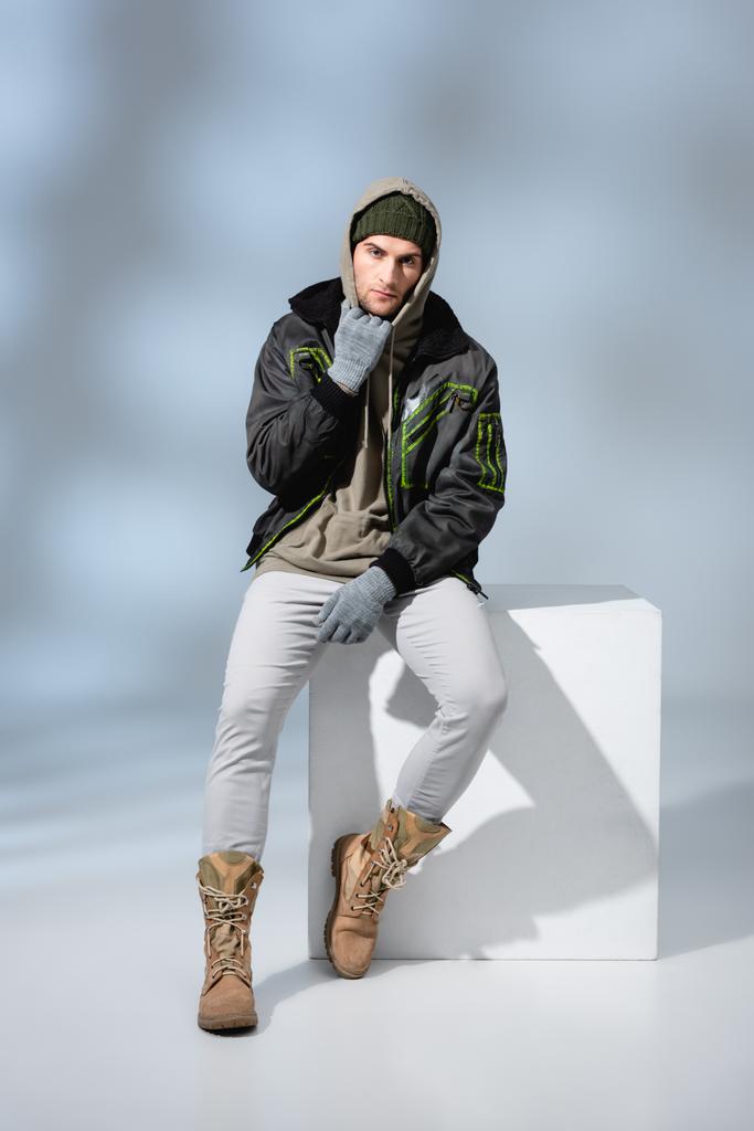 full length του trendy man σε καπέλο, γάντια και μπουφάν που ποζάρουν ενώ κάθονται σε λευκό κύβο σε γκρι  - Φωτογραφία, εικόνα
