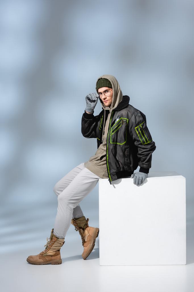 full length of trendy man in hat, gloves and parka adjusting glasses while sitting on white cube on grey  - Photo, Image