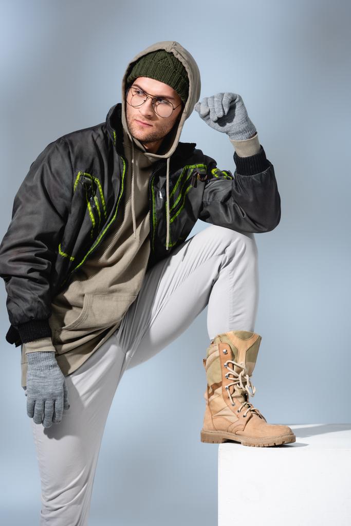 молодой модный человек в шляпе, перчатках и анораке опираясь на белый куб на сером  - Фото, изображение
