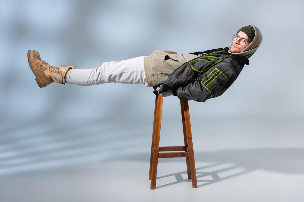 joven hombre de moda en sombrero y anorak acostado horizontalmente en taburete de madera en gris  - Foto, Imagen